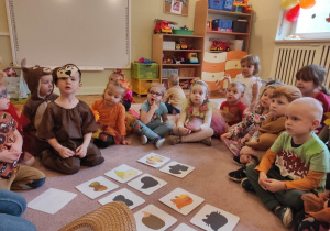 Dzieci przebrane w jesienne stroje siedzą w kole, pośrodku znajdują się ilustracje przedsztawiające jesienne motywy.
