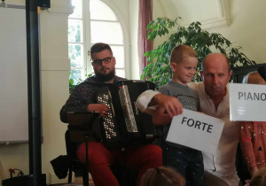 dwoje dzieci prezentuje kartki z napisami "Piano" "Forte". W tle mężczyzna grający na akordeonie.