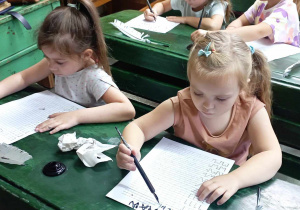Dwie dziewczynki z grupy 4 siedzą w ławkach i piszą piórem