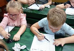 Dziewczynka i chłopiec z grupy 4 siedzą w ławkach i piszą piórem
