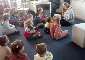 Dzieci z grupy 4 siedzą na dywanie i słuchają Pani z biblioteki czytającej książkę